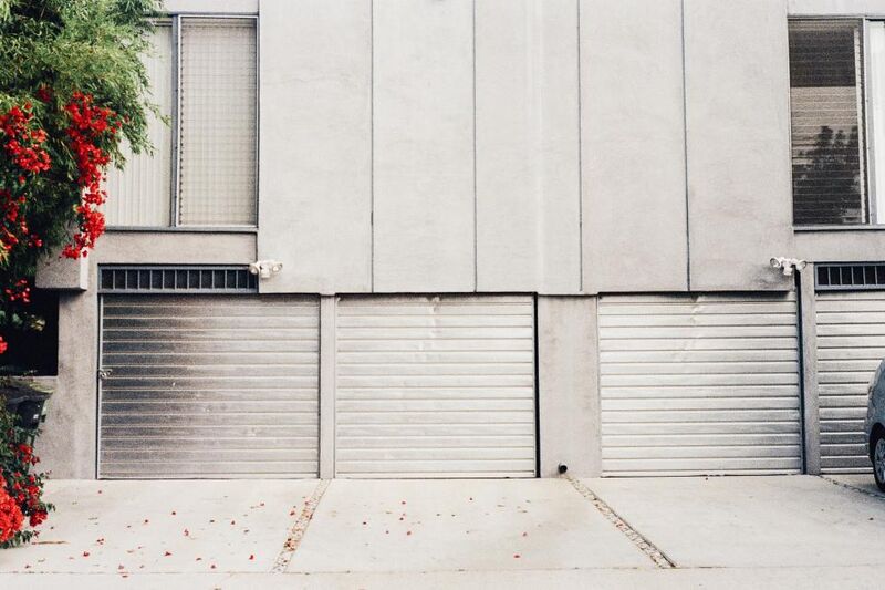 A suburban concrete driveway in front of twin roller-door garages with leaf litter blown across it